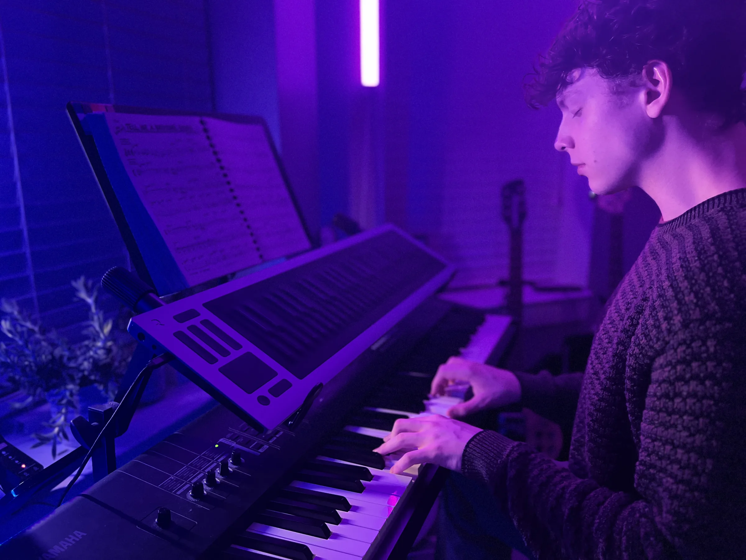 Nate Sage playing a keyboard in a studio lit violet, a chart on the music stand